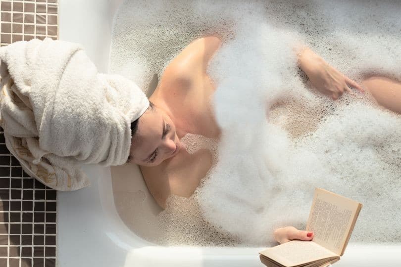 A woman with her hair in a towel, reading a book while taking a hot bubble bath
